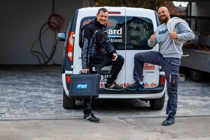 Zwei Elektriker mit Messkoffer und Kabelring am Servicefahrzeug